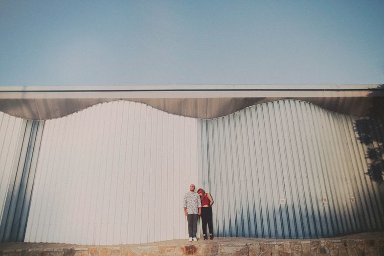 A couple stands against a white corrugated metal wall in an industrial setting.