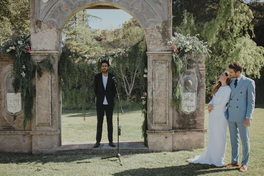 Wedding party members posed beneath a decorated stone archway in a garden setting.