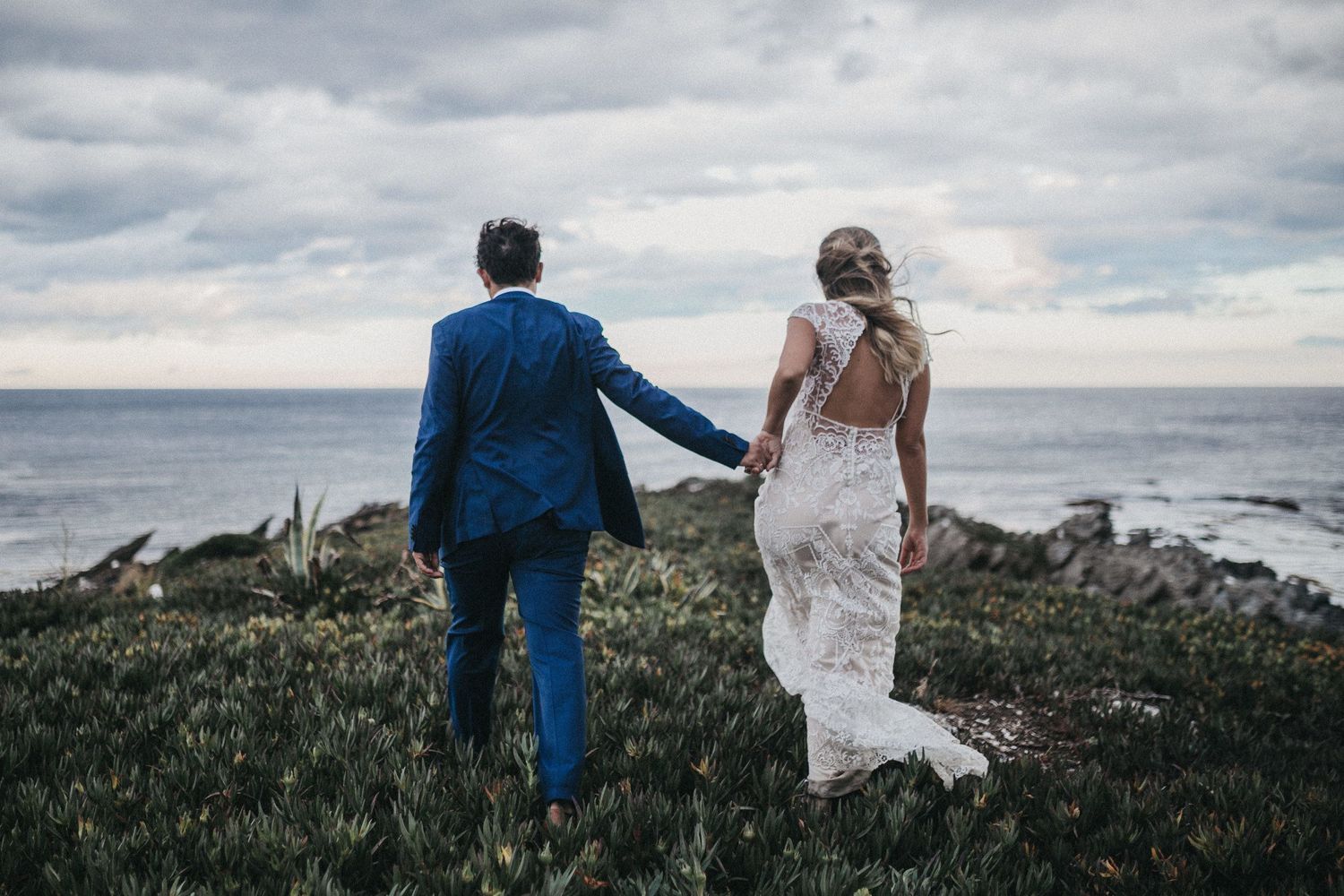 A couple in wedding attire holds hands while standing on a cliff overlooking the ocean under moody skies.
