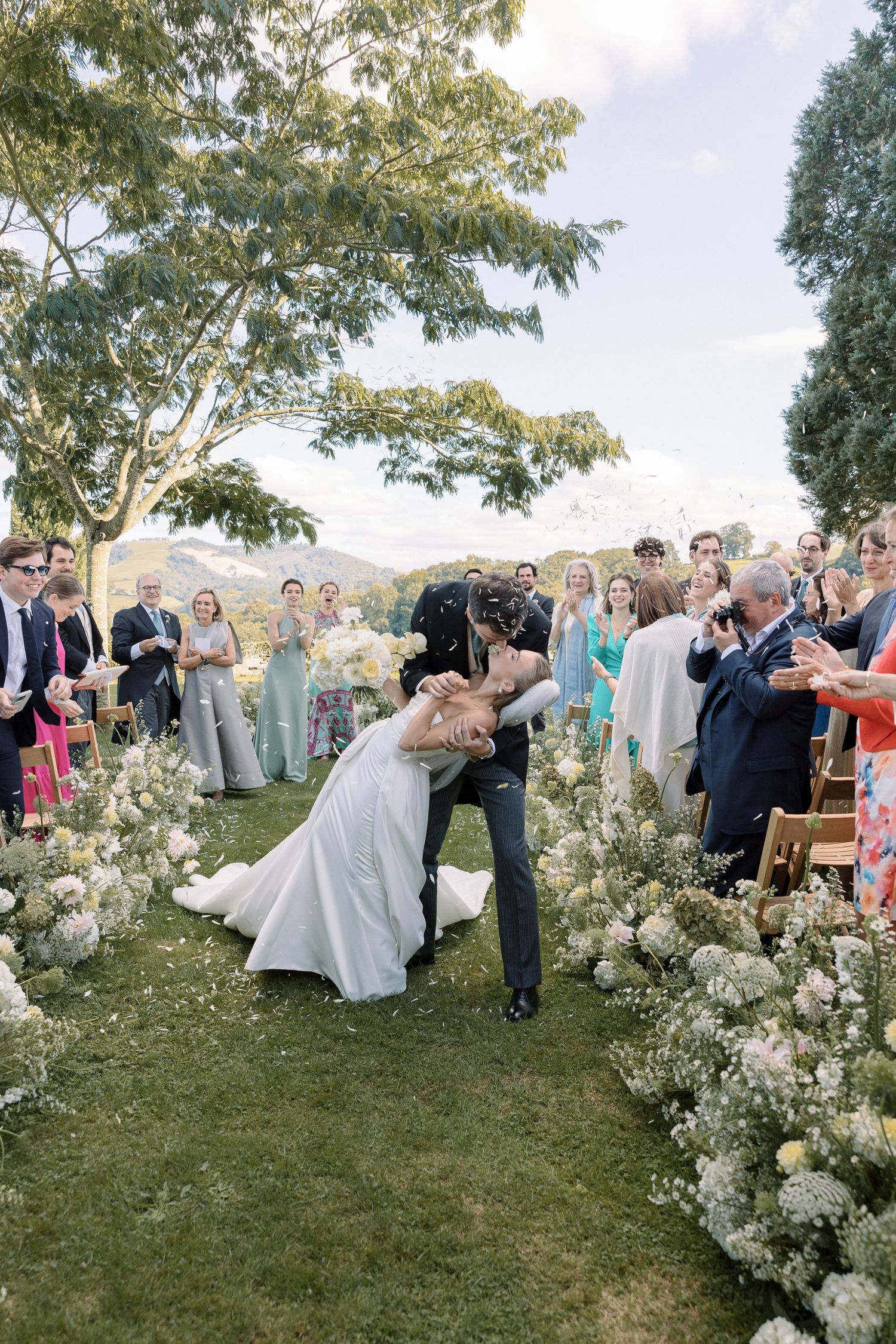 Bride and groom share a romantic dip kiss while walking down a flower-lined aisle at an outdoor wedding ceremony.