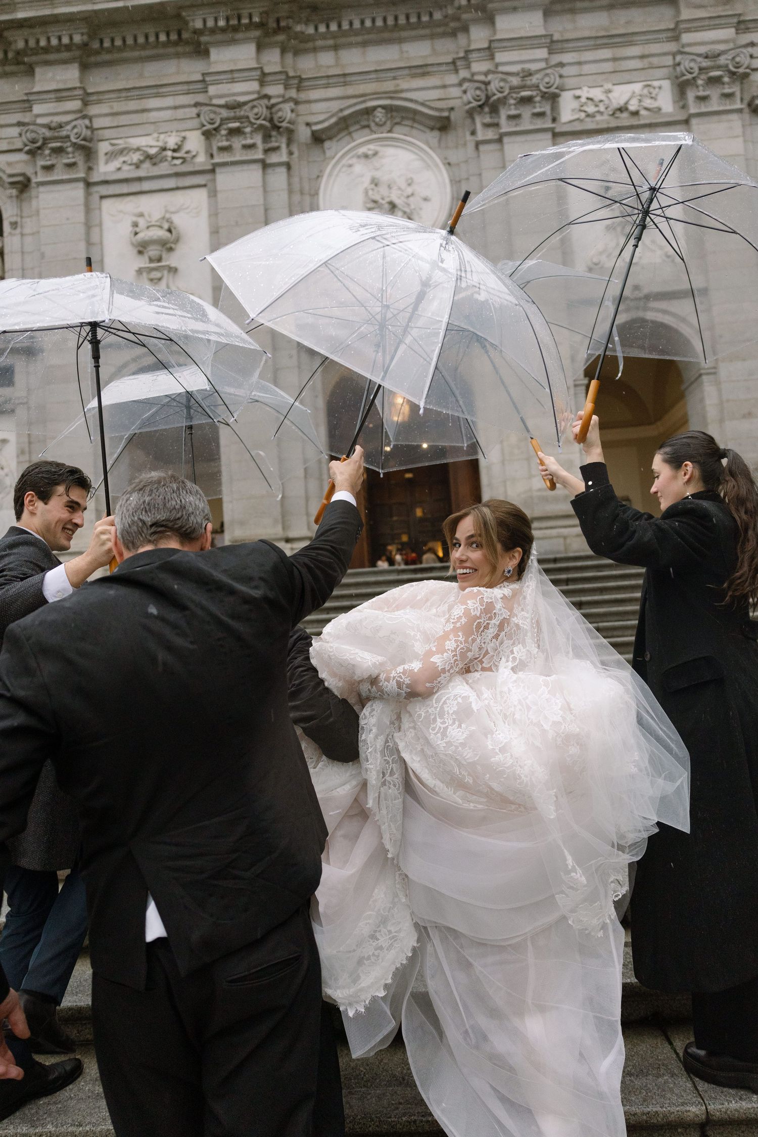 Bride in white lace gown smiles surrounded by guests holding clear umbrellas outside an ornate church on a rainy wedding day.