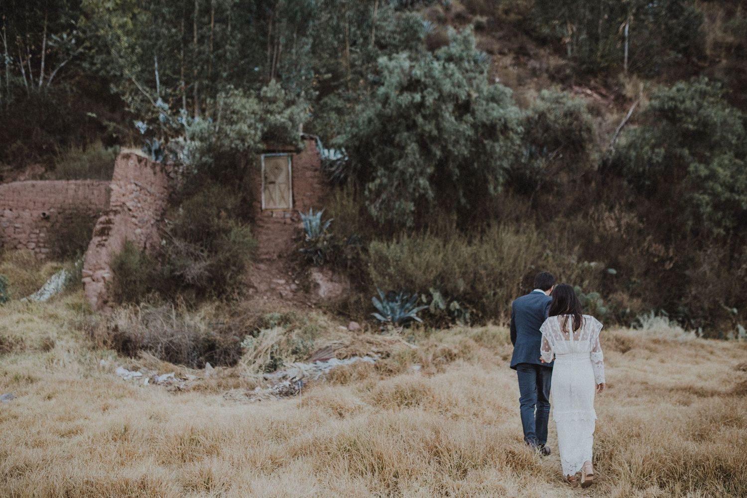 A couple stands together in wedding attire against a backdrop of ancient stone ruins and lush foliage.