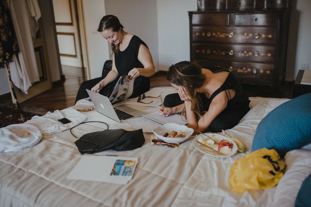 Two people sitting on a bed wearing black clothes while looking at paperwork and laptops surrounded by documents.