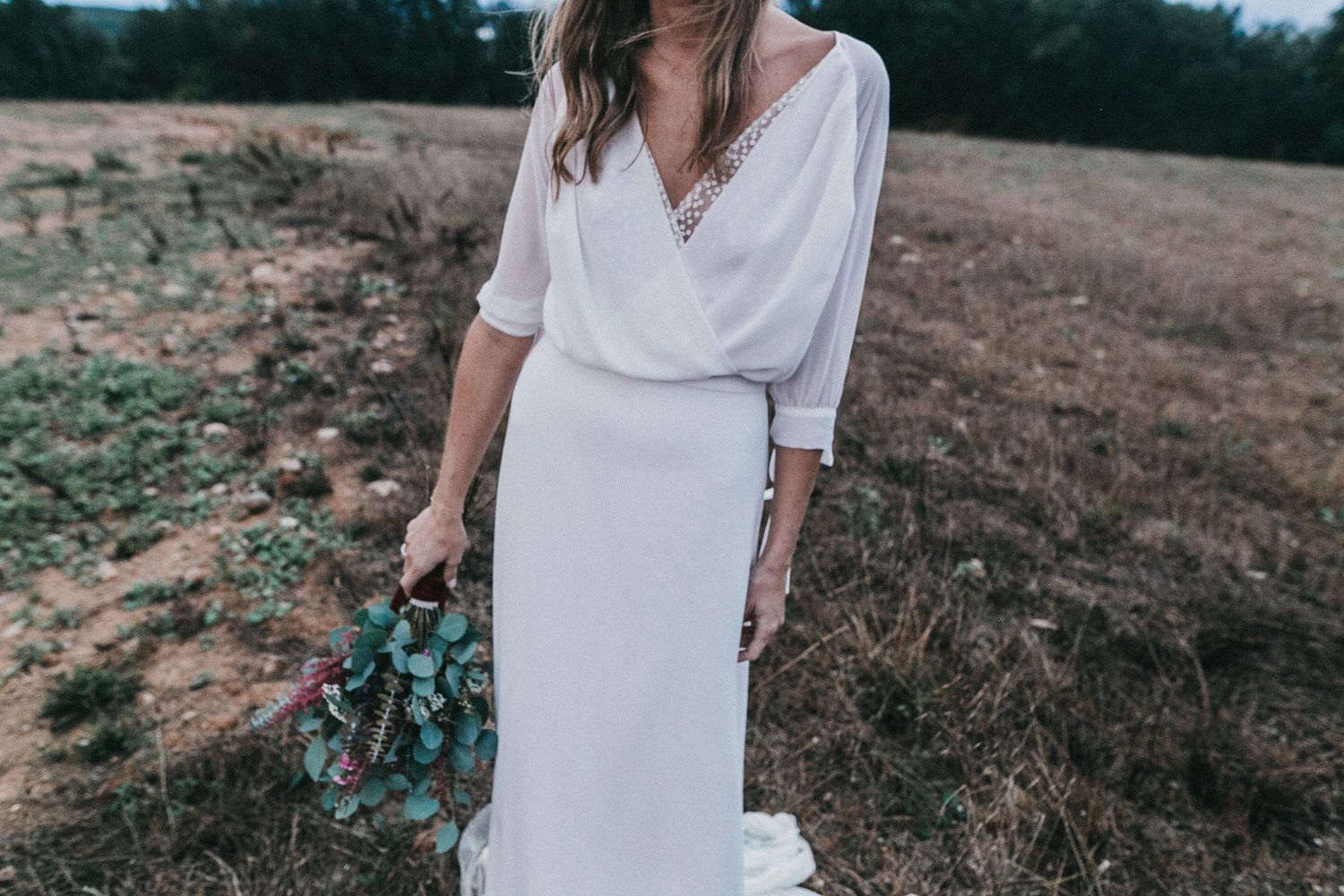 Bride in flowing white dress holding a teal bouquet in rustic outdoor setting.
