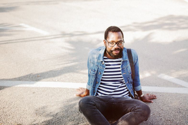 A person in striped shirt and denim jacket sits cross-legged on pavement.