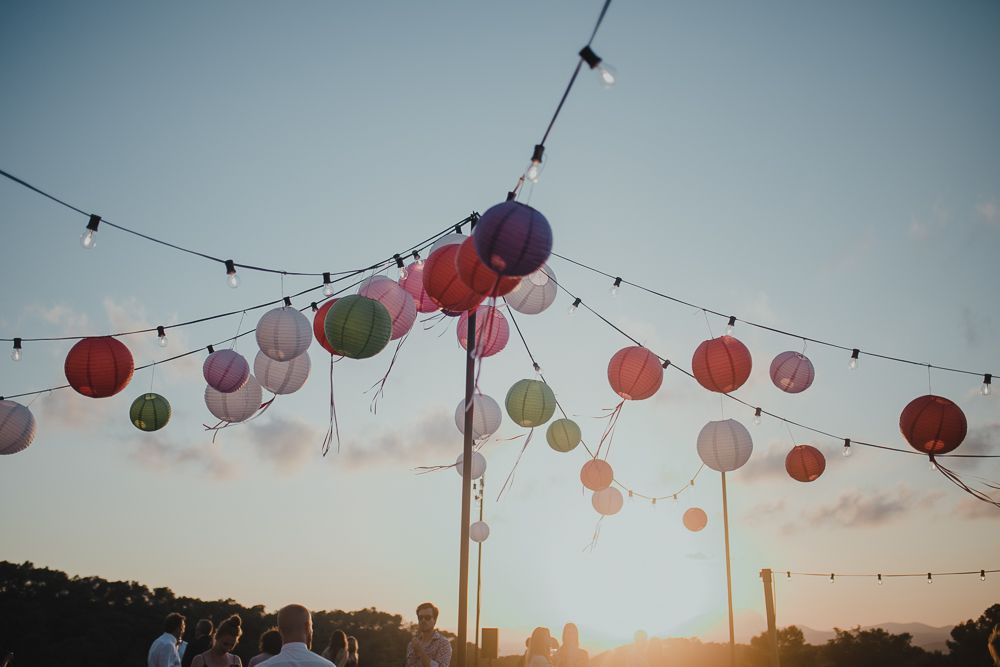 String lights adorned with colorful paper lanterns hang against a sunset sky at an outdoor celebration.