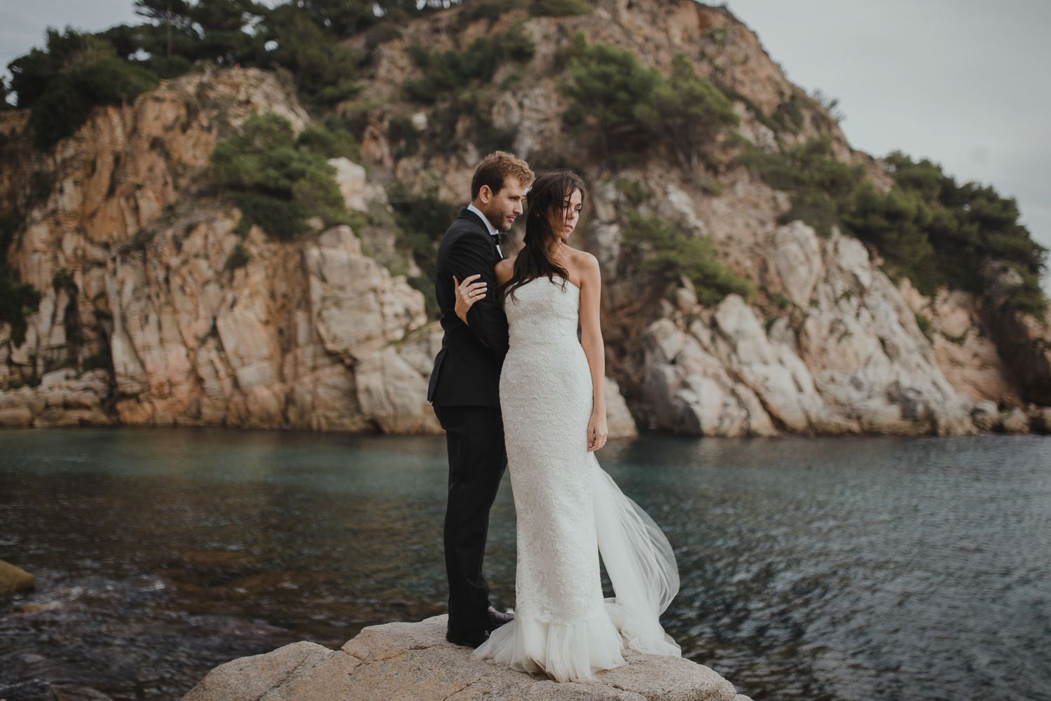 A couple in wedding attire standing on coastal rocks with dramatic cliffs in the background.