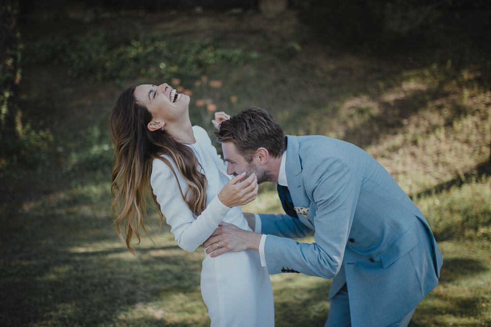 Couple in wedding attire shares playful candid moment while laughing together outdoors.