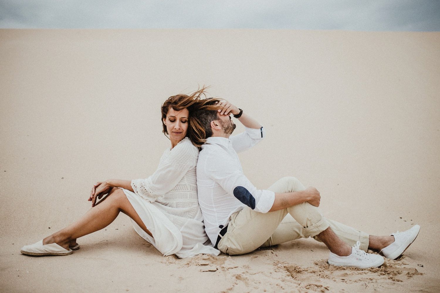 Playful couple in white outfits sitting back to back on sandy beach.