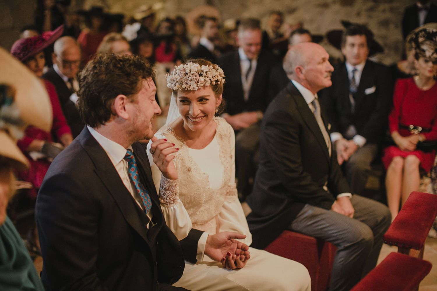 Intimate wedding ceremony in rustic stone church with guests seated on red chairs.