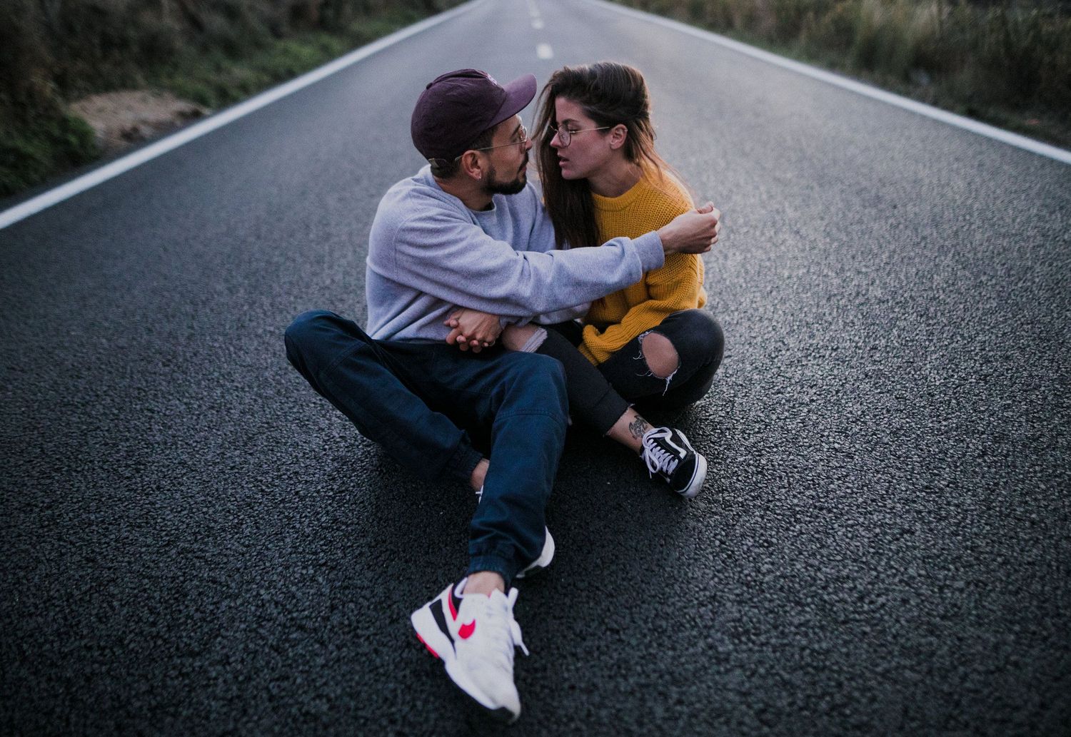 A couple sitting intimately on an empty road wearing casual attire.