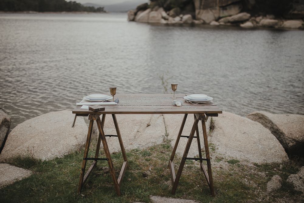 Empty rustic wooden table set up for dining on rocks by a serene lake.