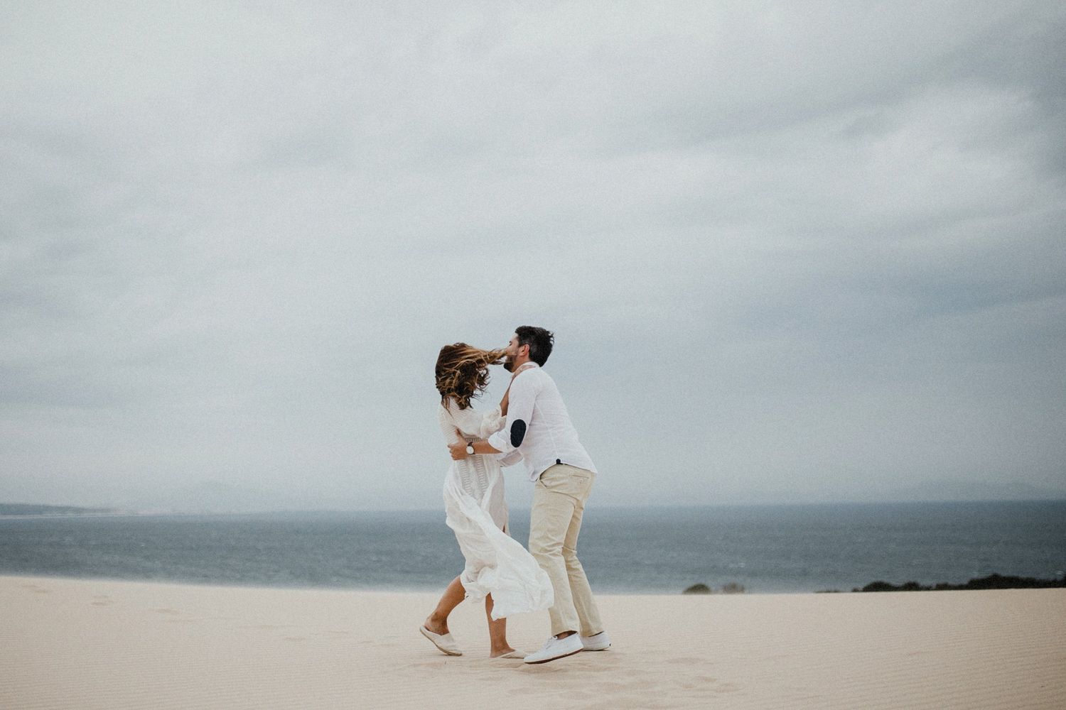 Two people dancing together on a beach with a moody grey sky overhead.