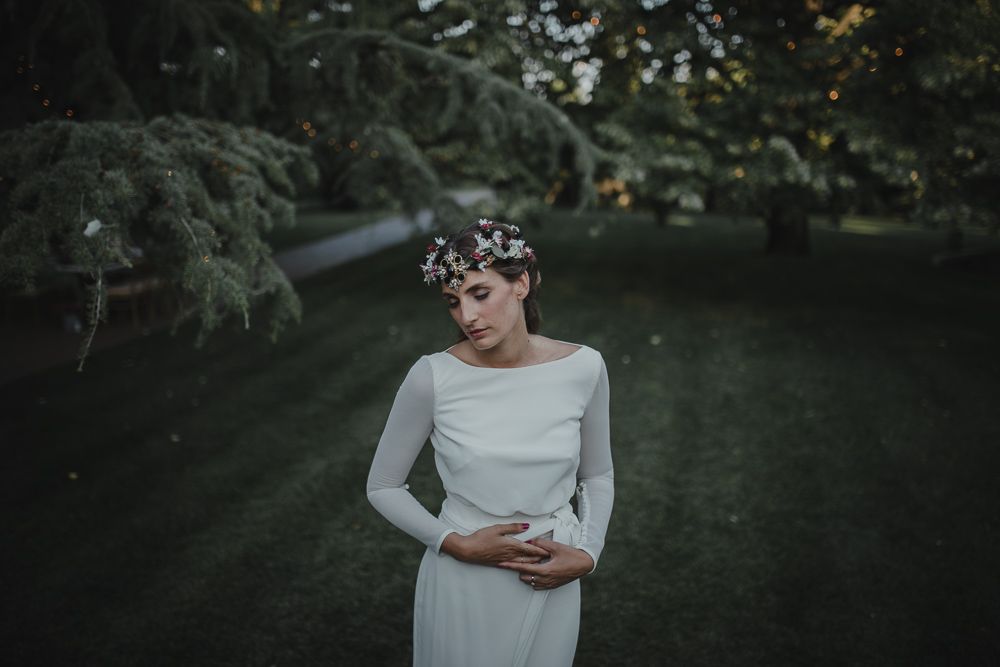 A bride in a simple long-sleeved dress poses in a garden setting with a floral crown.