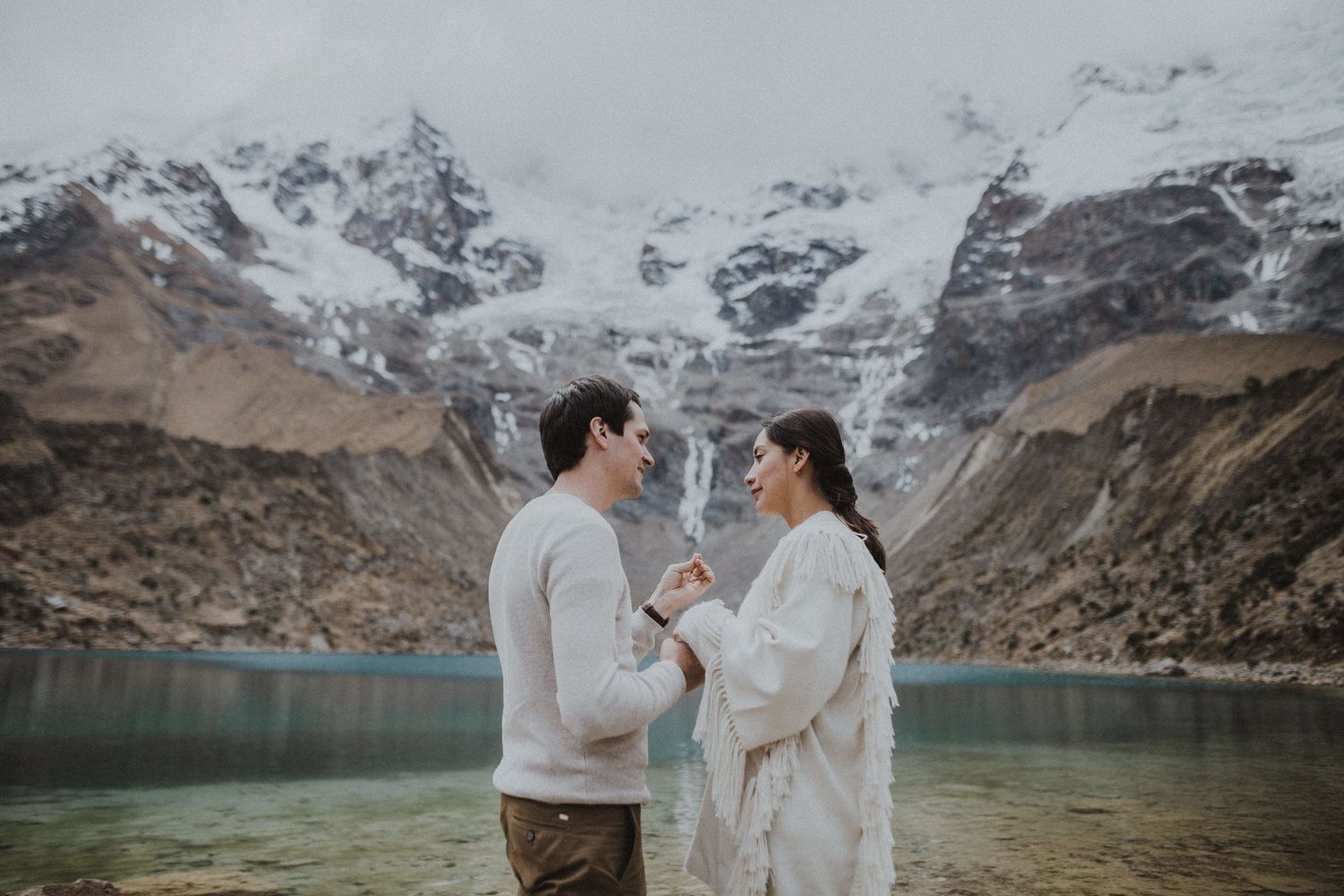 A couple shares a tender moment beside a pristine mountain lake with snowy peaks in the background.
