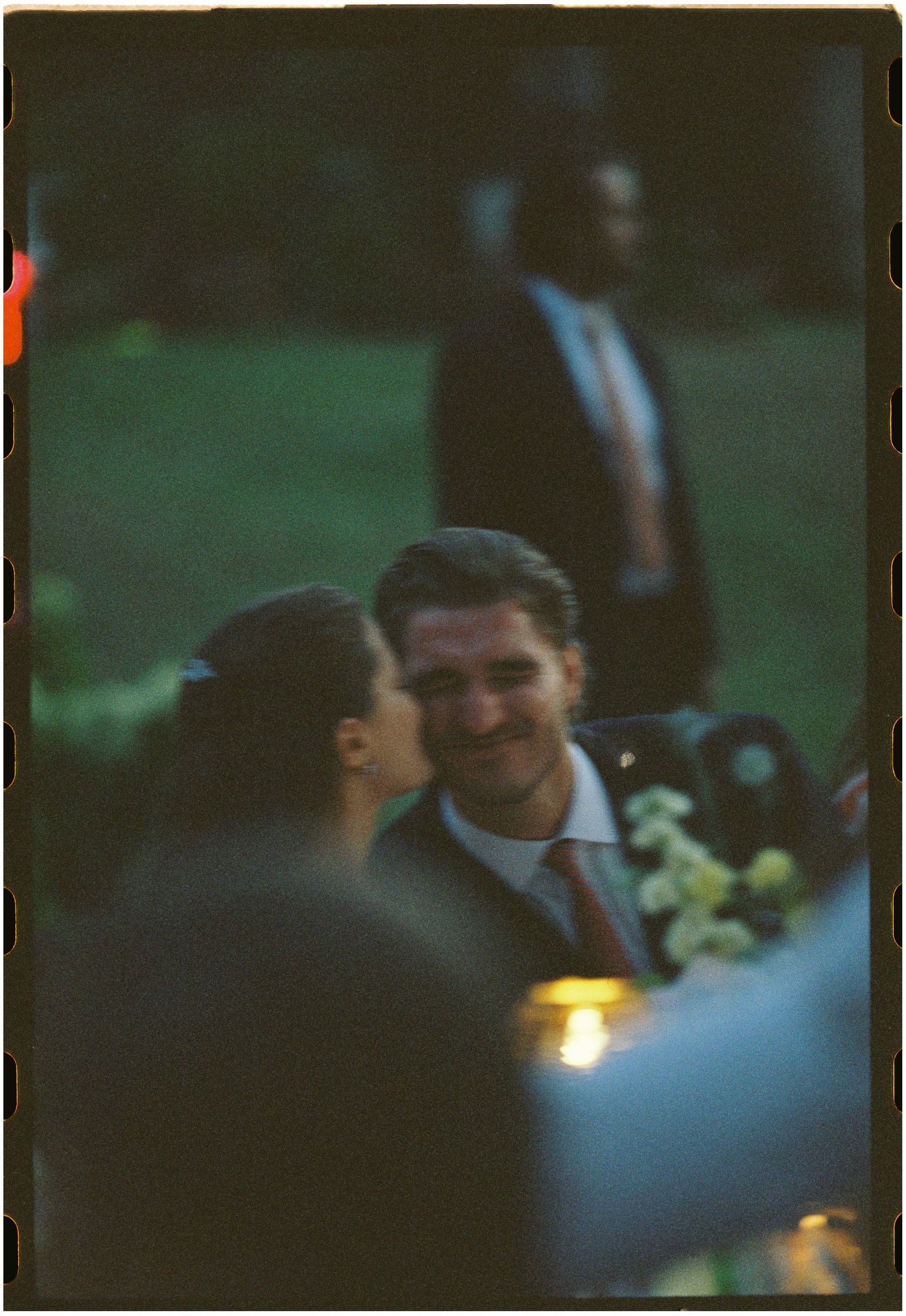 A couple shares a romantic moment at an evening outdoor event, with a man smiling as a woman leans in close.