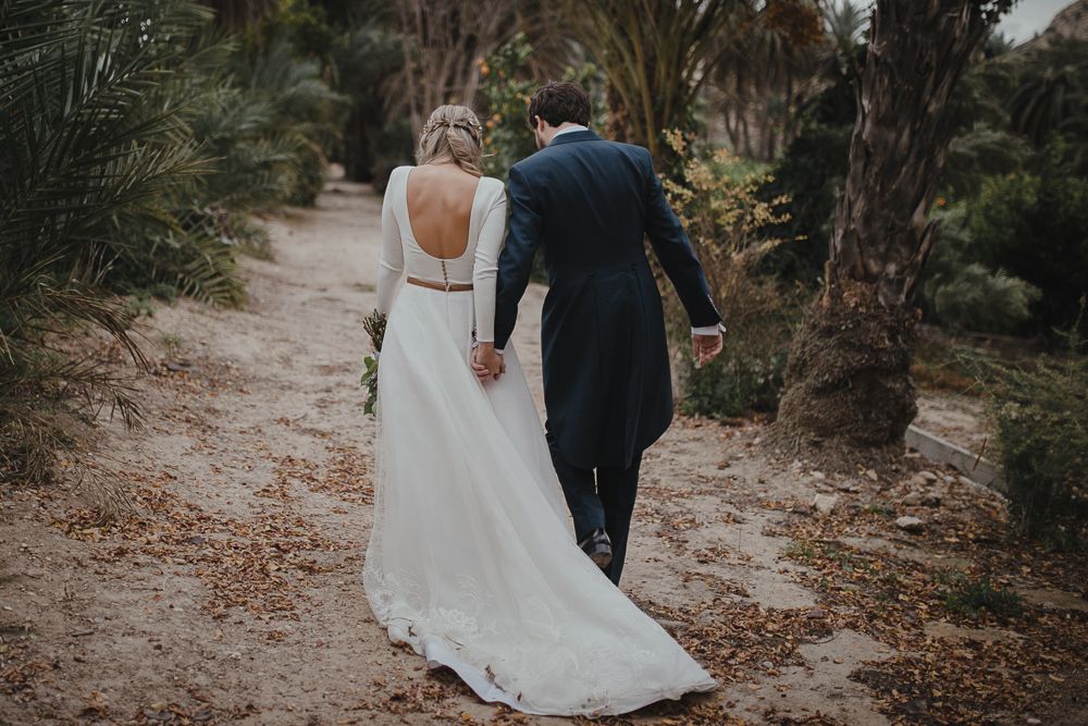 A couple in wedding attire walks down a palm-lined path holding hands.