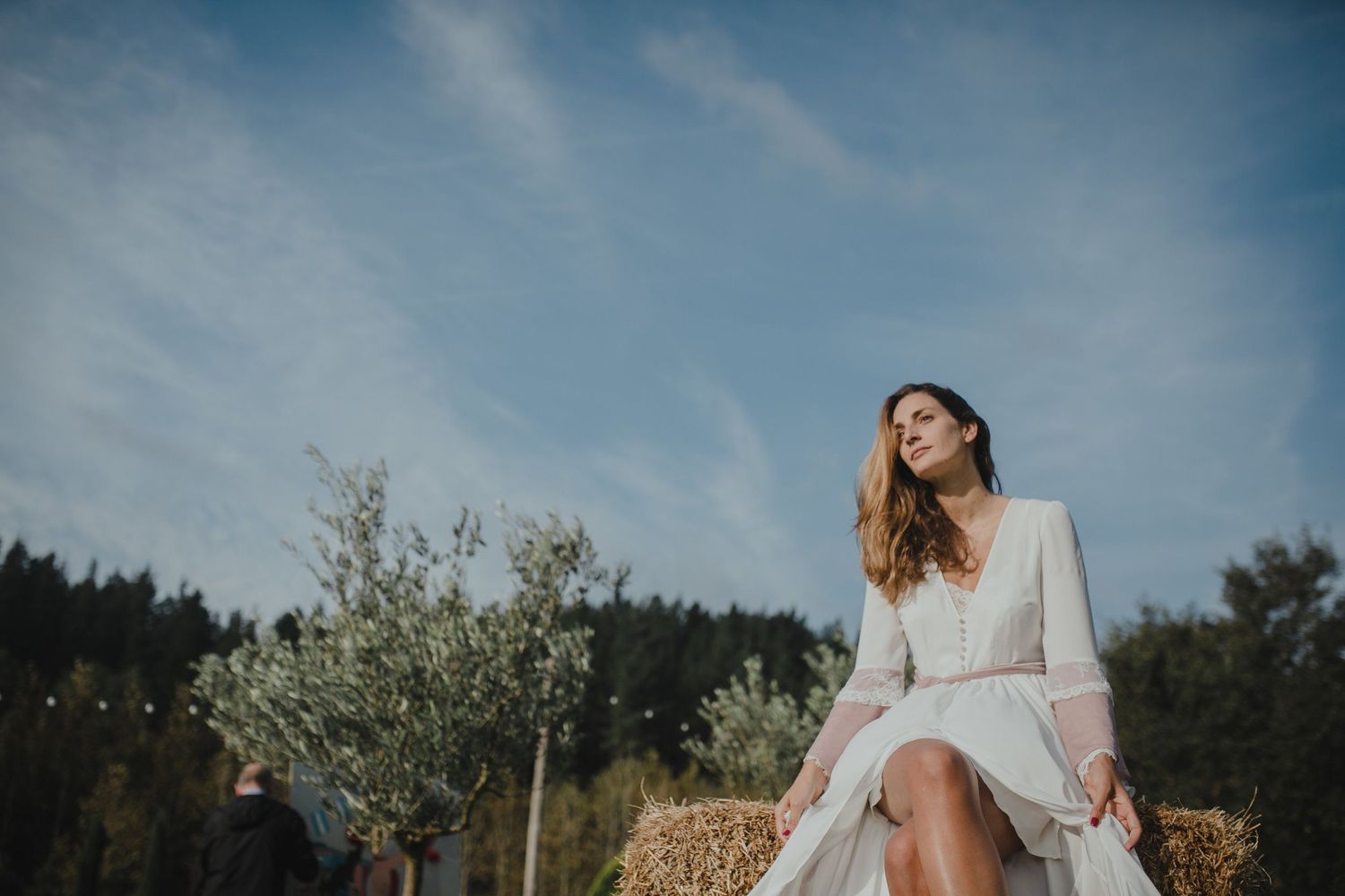 Someone in a flowing white dress sits dreamily on a hay bale against a nature backdrop with cloudy skies.