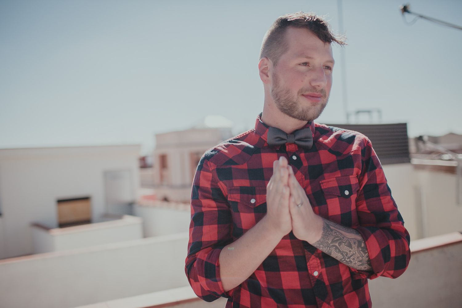 Person in red plaid shirt with tattoos stands in prayer pose on urban rooftop.