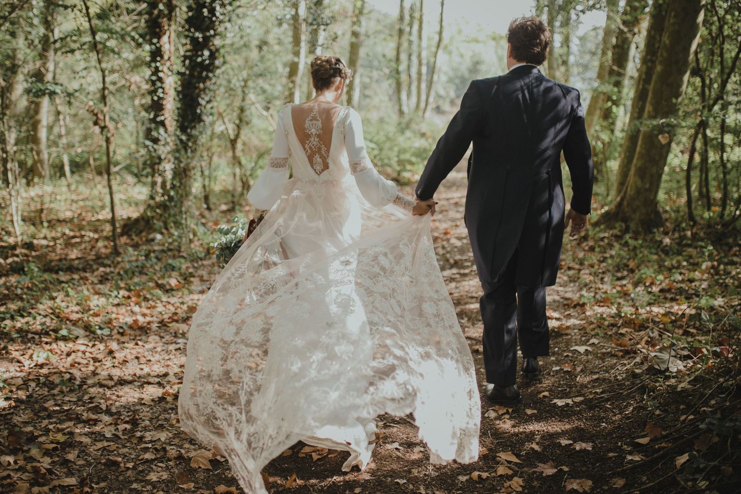 A couple walking hand in hand through a wooded path, bride's dress flowing behind them.