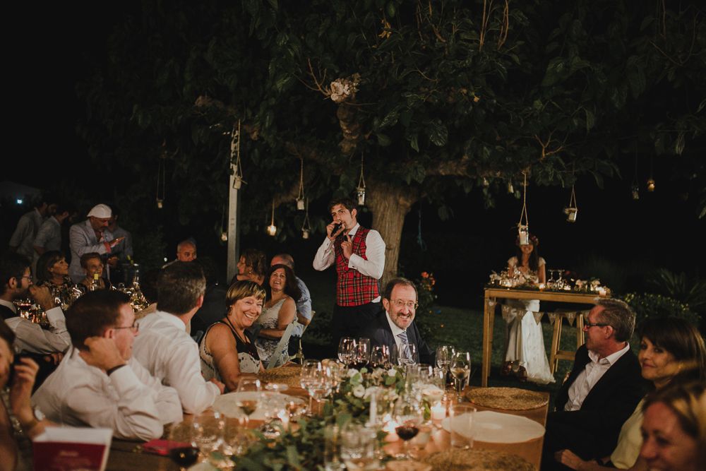 Evening outdoor reception with guests gathered at decorated tables under string lights.