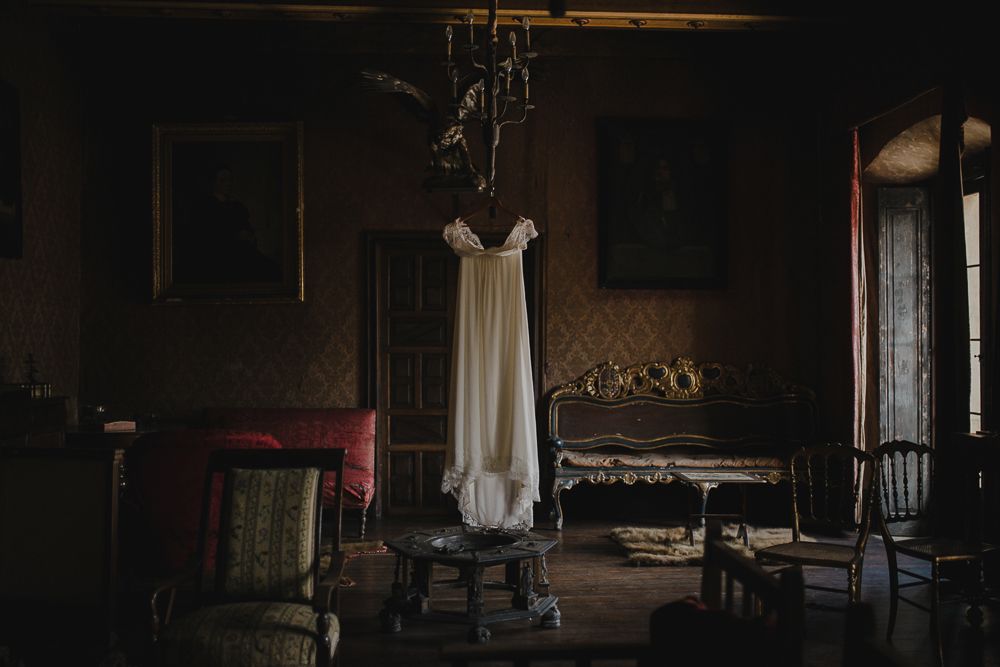 White wedding dress hanging from chandelier in a dark vintage room with antique furniture.
