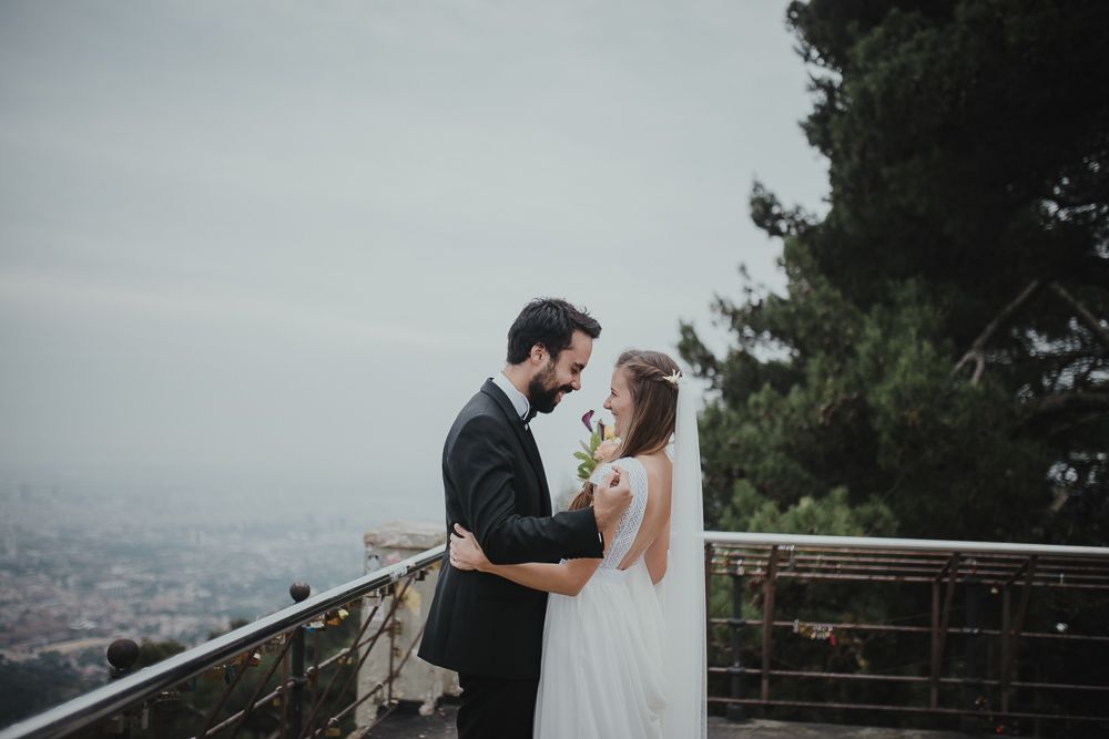 A romantic wedding couple embraces on a scenic overlook with city views below.