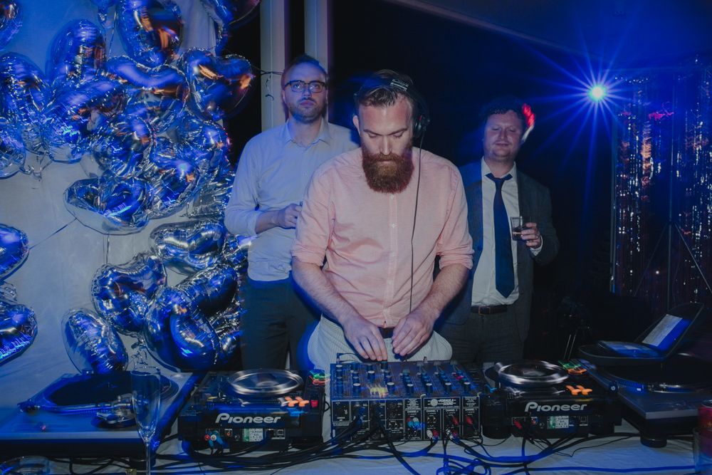 DJ in pink sweater mixing music at a party with blue lighting and decorative backdrop.