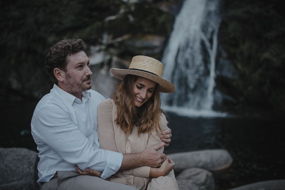A couple shares an intimate moment by a waterfall, with one wearing a straw hat.