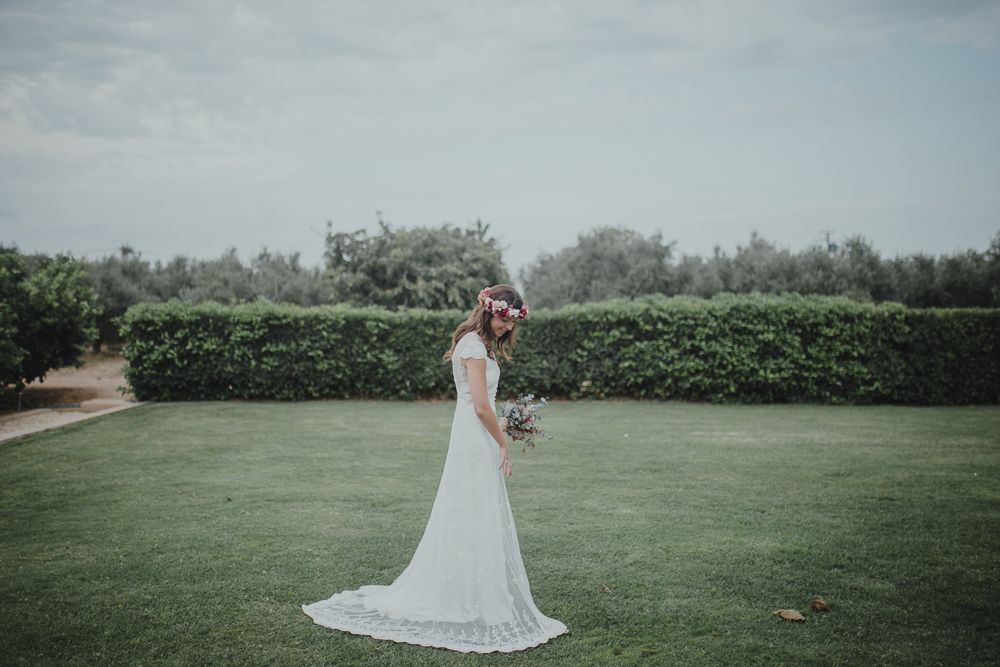 A bride in a flowing white dress stands on grass holding a floral bouquet.
