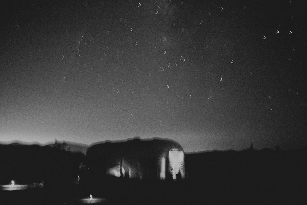 Black and white nighttime landscape with silhouetted buildings and star trails in the sky.