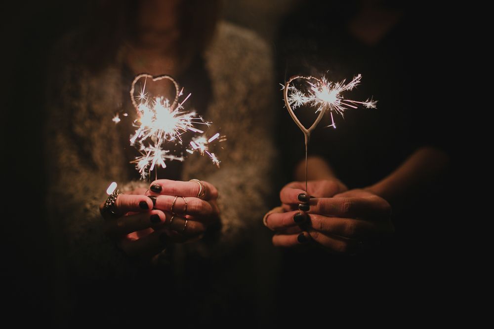 Two hands holding lit sparklers against dark background.