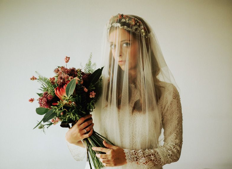 Ethereal bridal portrait with sheer veil, floral crown, and vibrant bouquet against cream background.