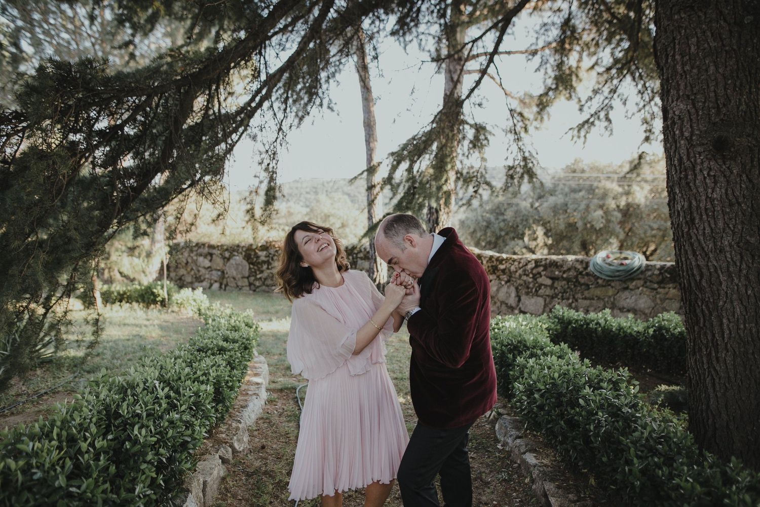 A couple shares a romantic moment under pine trees in a garden setting.