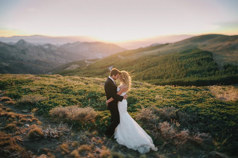A couple in wedding attire embraces on a hilltop during golden hour with mountains in background.
