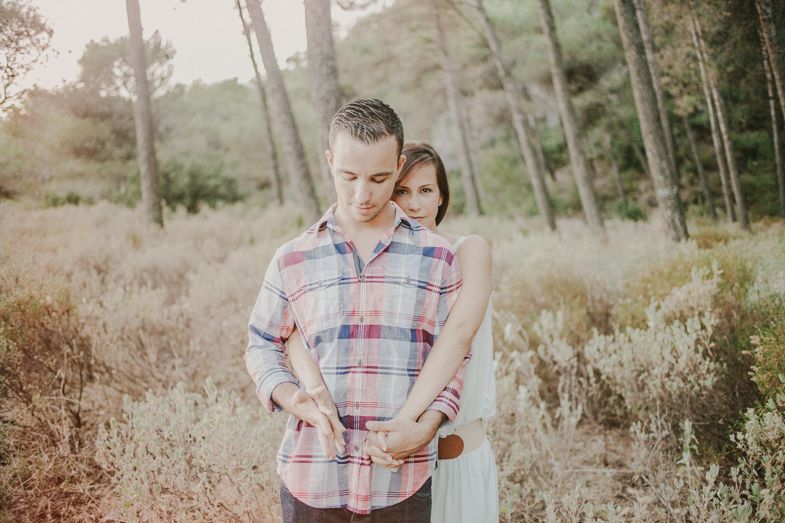 A couple embraces in a sunlit forest clearing wearing casual attire.