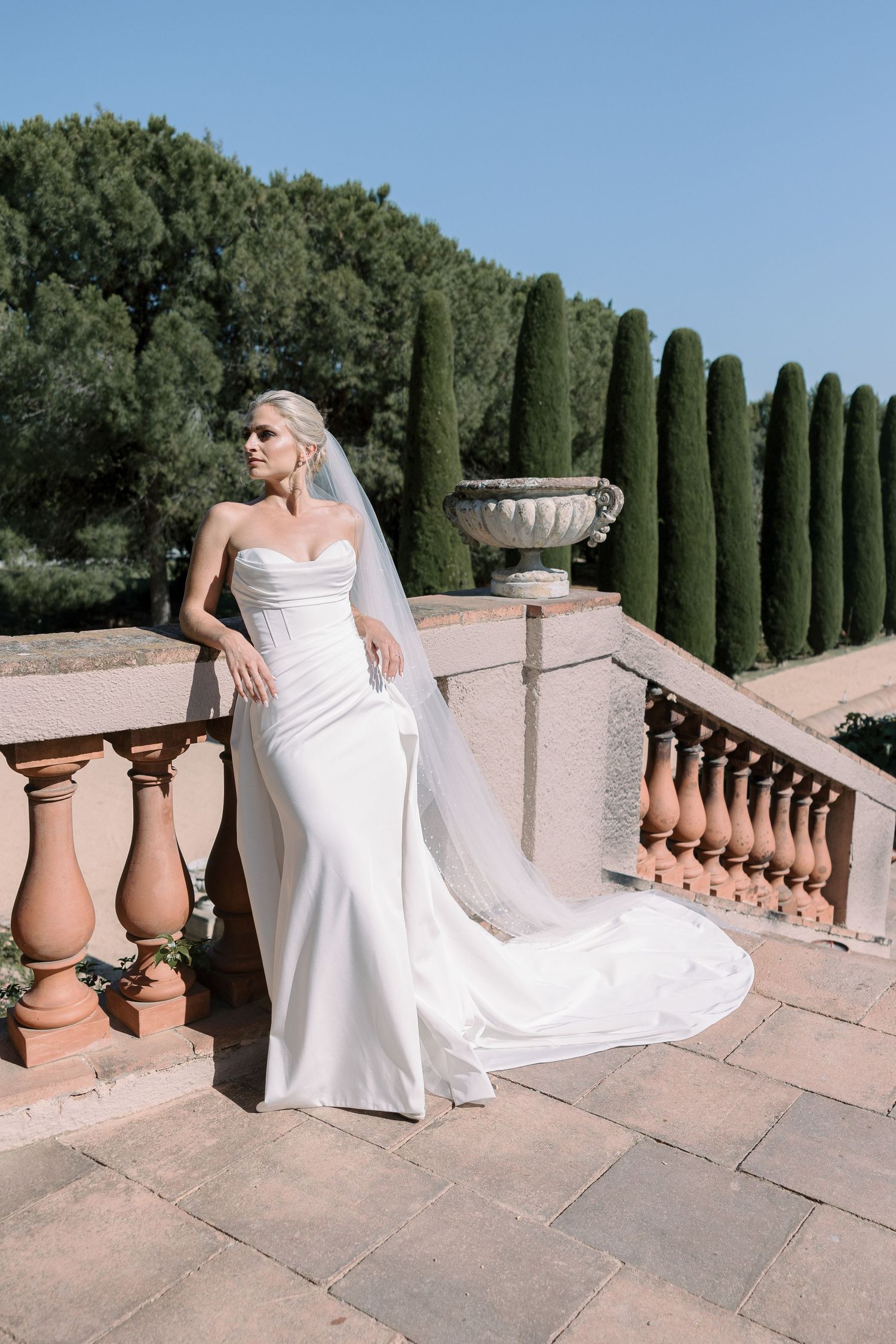 Elegant bride in fitted white gown posing by stone balustrade with cypress trees.