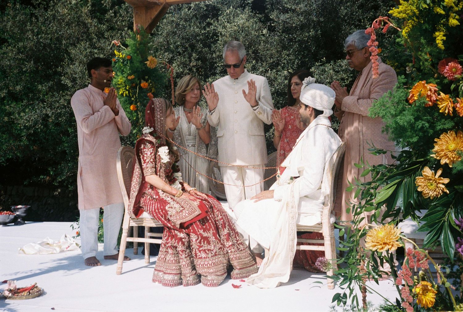 A wedding ceremony takes place outdoors with guests gathered around a white-draped altar surrounded by yellow flowers.