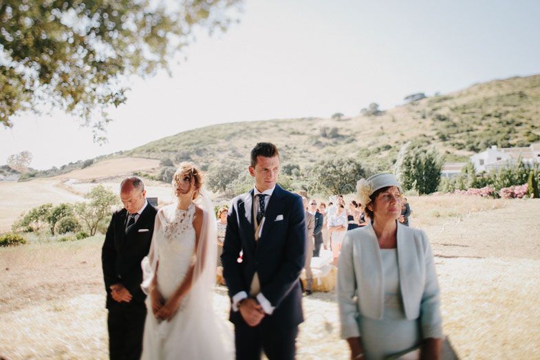 A wedding party stands in a line during an outdoor ceremony on a sunny hillside.