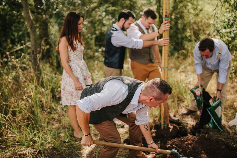 Casual wedding party helps plant a tree together in a sunny garden setting.