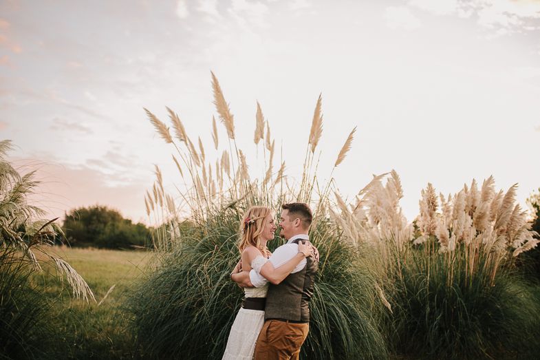 A couple embraces in front of tall pampas grass during a dreamy sunset photoshoot.