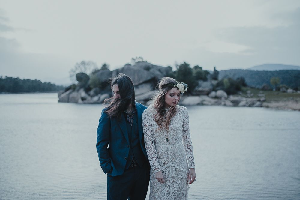Moody wedding portrait by rocky lake shore with mountains in background.