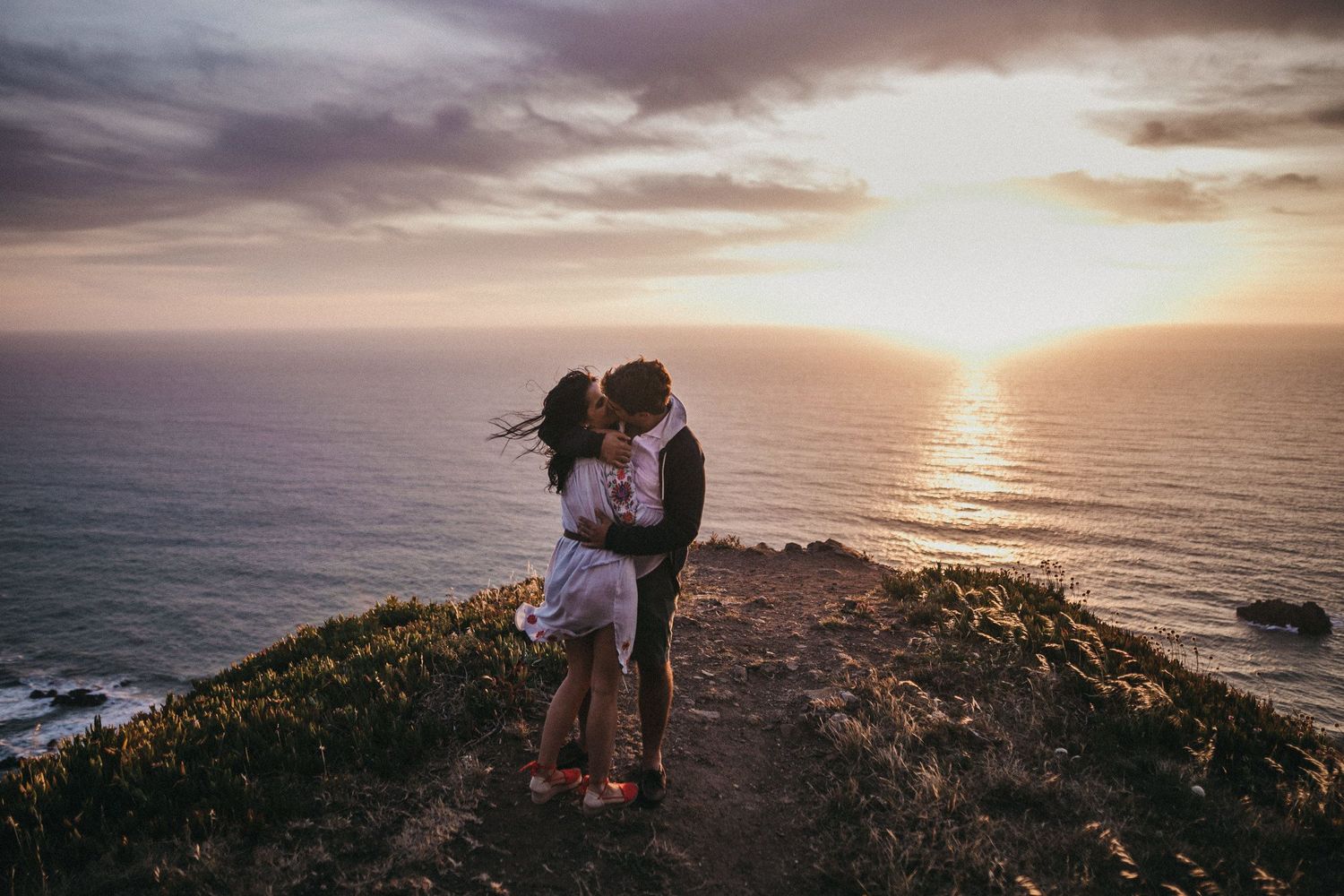 Couple embracing on a coastal cliff during golden hour sunset over the ocean.