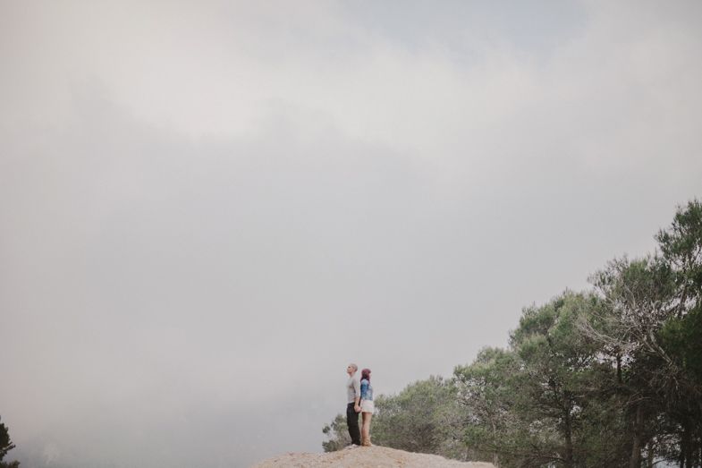 Two silhouettes standing together on a rocky overlook against a foggy sky.