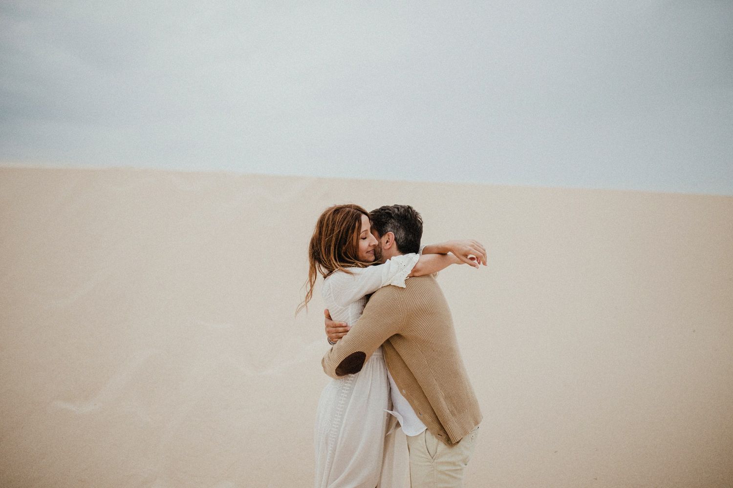 Two people share an intimate embrace while standing in a desert setting with soft lighting.