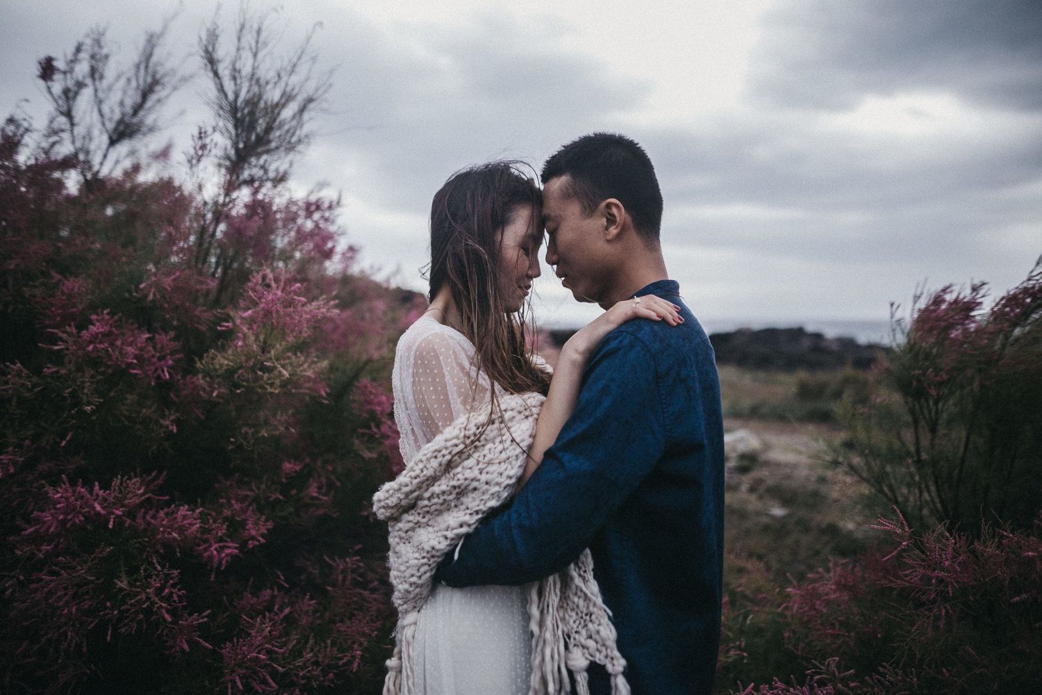 Couple embracing among pink flowering bushes under cloudy sky.