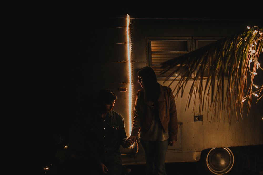 Two people stand near a vintage trailer with light effects at night.