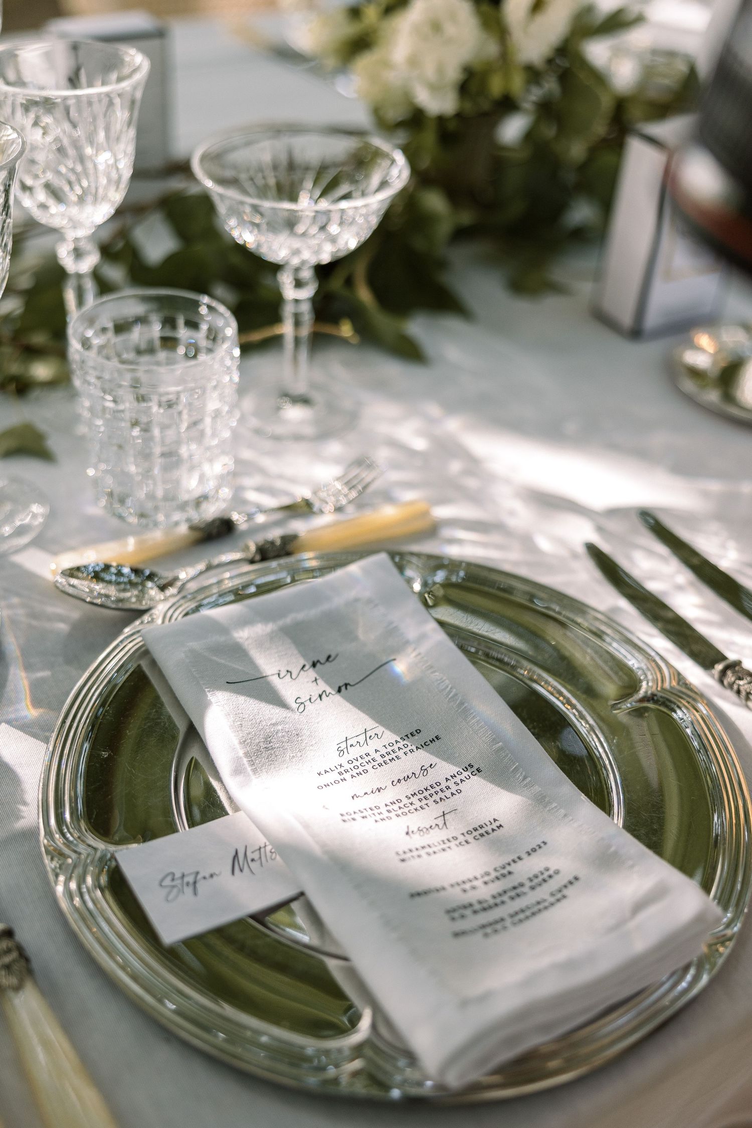 Elegant wedding reception place setting with crystal glasses and menu card on silver charger plate.