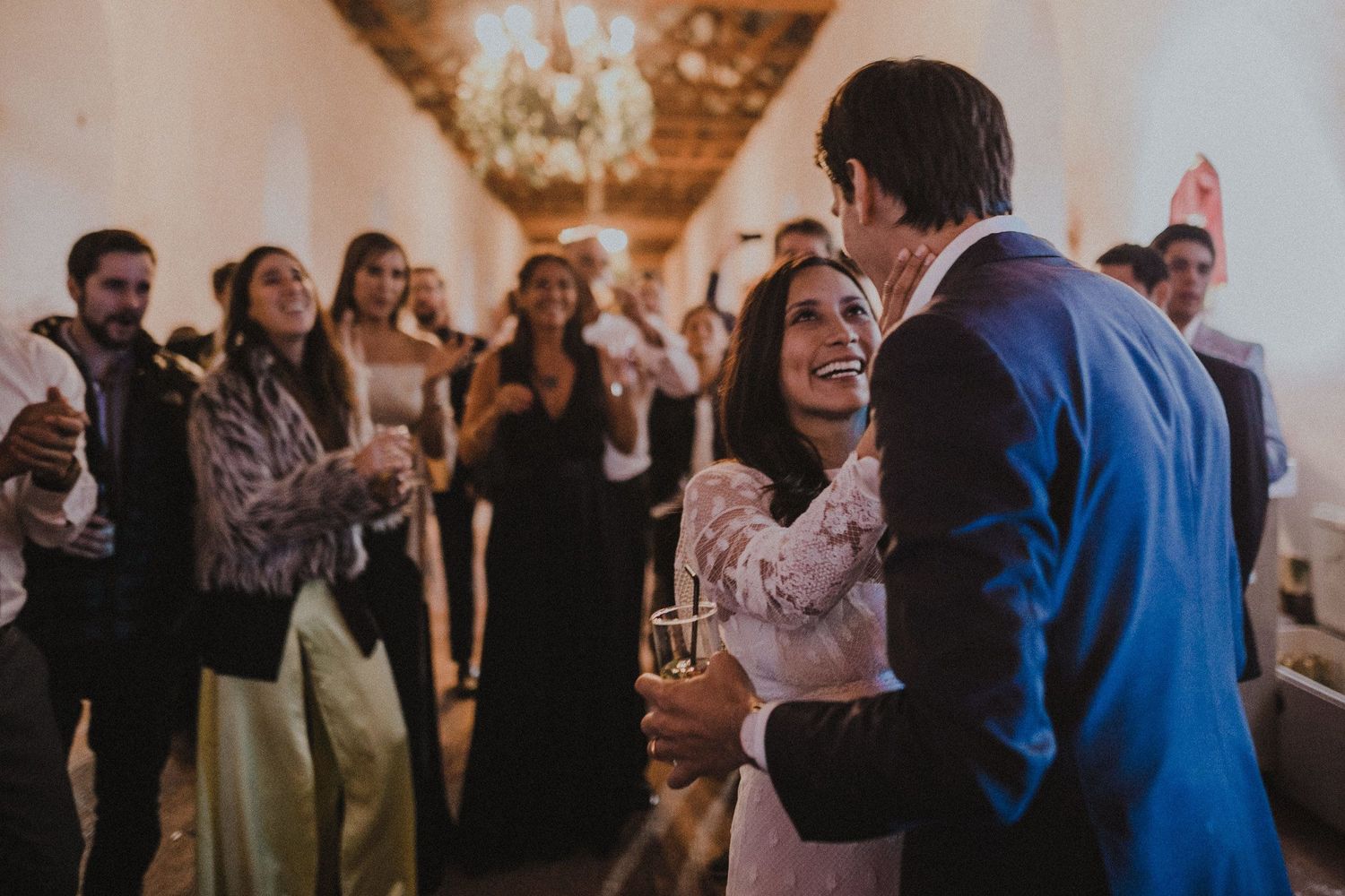 Couple sharing intimate moment while guests celebrate in rustic indoor venue with chandelier.