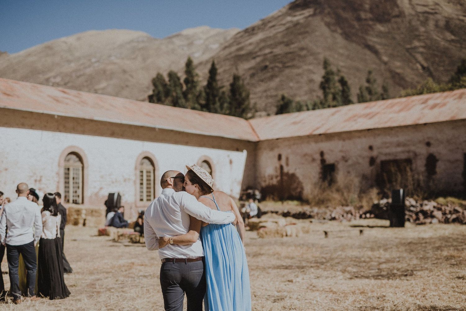 Guests embrace at a rustic outdoor wedding celebration with stone buildings and mountains in the background.